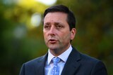 Victorian Opposition Leader Matthew Guy, who is wearing a suit. The background behind him is blurry and green.