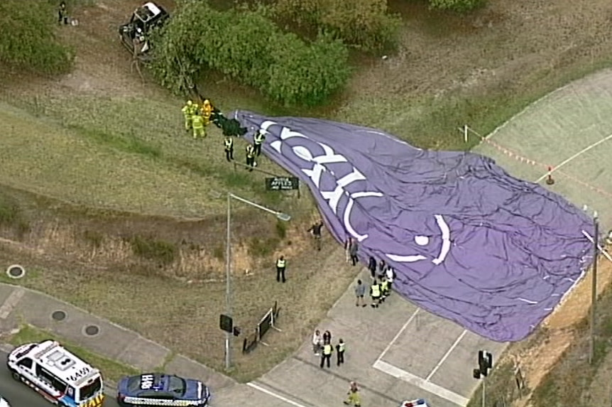 A purple hot air balloon lies deflated and laid out on a footpath next to a busy intersection.
