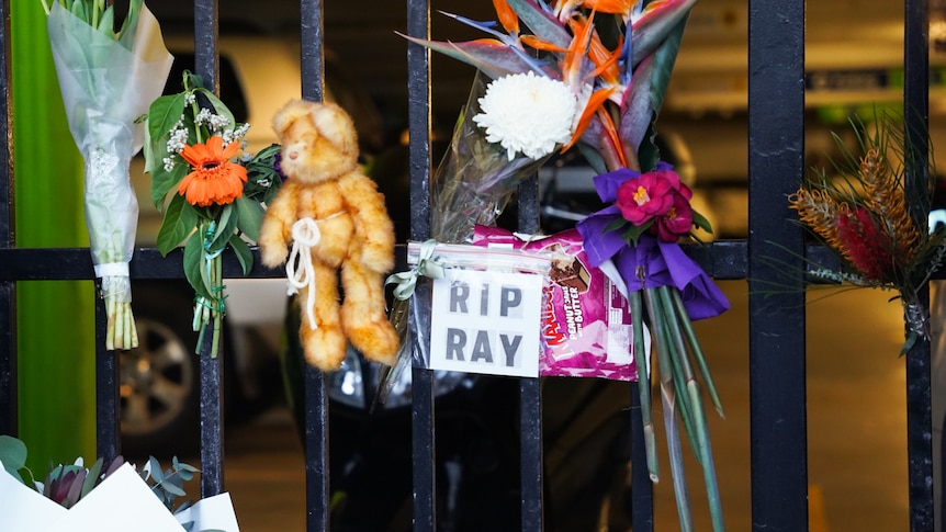 Image of flowers, teddy bear sign reading 'RIP Ray' and  Maxibon ice cream box strapped to fence