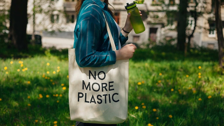 A woman holding a reusable cotton zero waste bag with text No More Plastic, while drinking from a plastic water bottle.