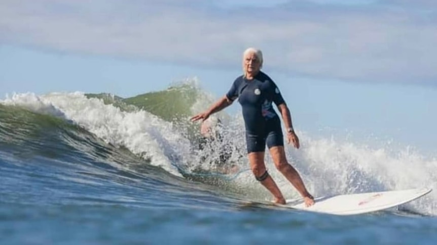 A woman with white hair and a dark wetsuit riding a wave on a surfboard