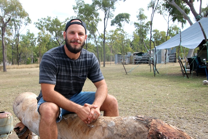 A man with a beard and a baseball cap on backwards sits on a log at a camp