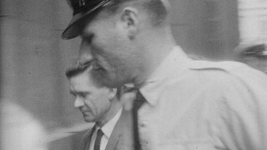A historical black-and-white image of a police officer escorting a well-dressed criminal into a courthouse through a crowd
