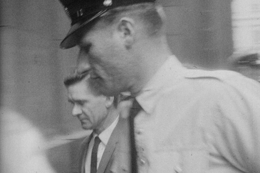 A historical black-and-white image of a police officer escorting a well-dressed criminal into a courthouse through a crowd