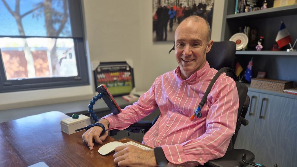 Smiling bald man, wears pink, white striped shirt, has an ear speaker, a breathing tube over shoulder, phone strapped to hand.