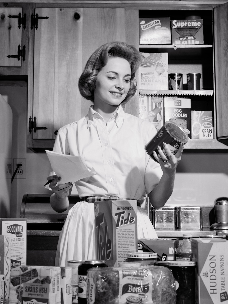 A black and white image of a smiling white woman in 1960s dress, looking at a can.