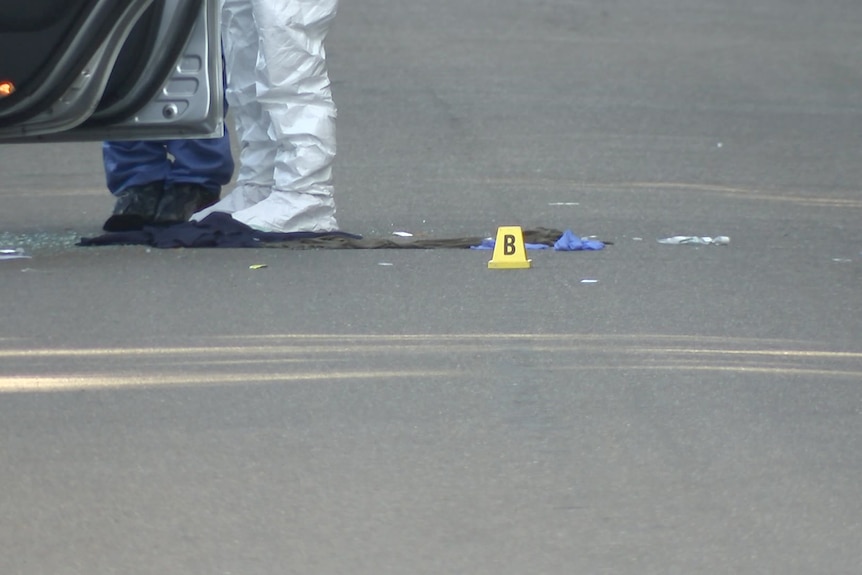 a forensic officer stands outside a car after a shooting with a number marker placed on the floor