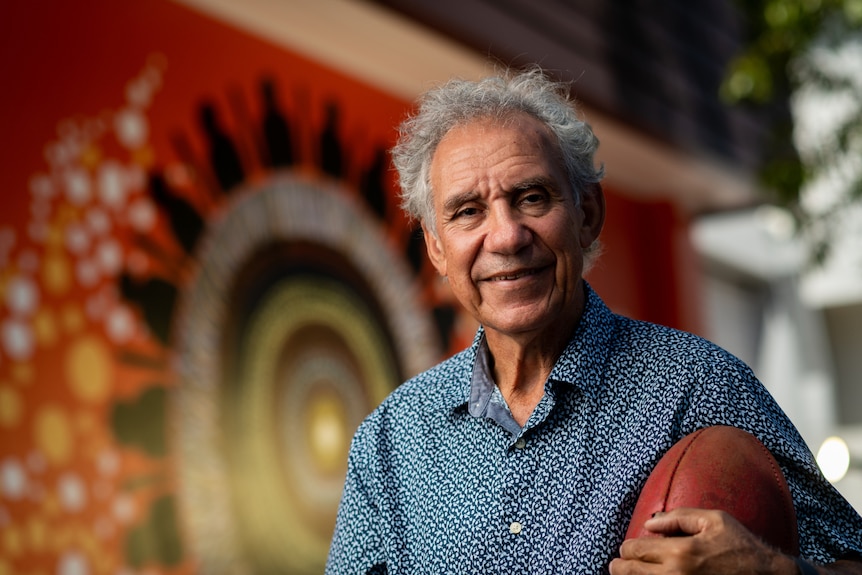 A smiling man holding a football with a mural in the background. 
