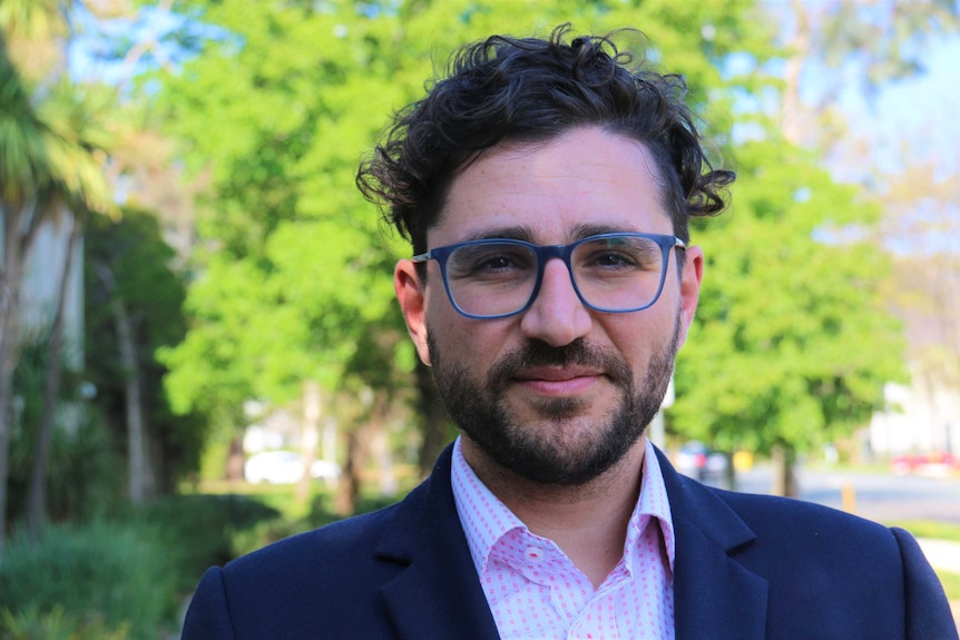 A man in a suit, with curly dark hair and glasses, green trees behind. 