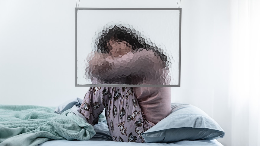 A girl sits up in bed, hugging her knees to her chest. Her face is blurred by a sheet of tempered glass.