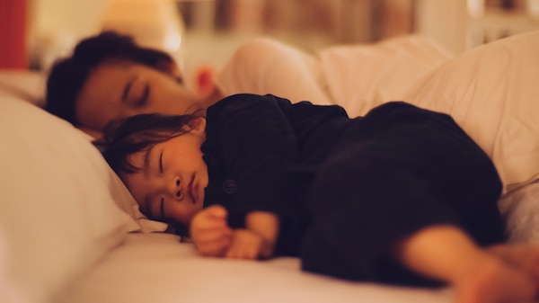 A young child sleeps and a sleeping mother is just visible behind them, in a room dimly lit.
