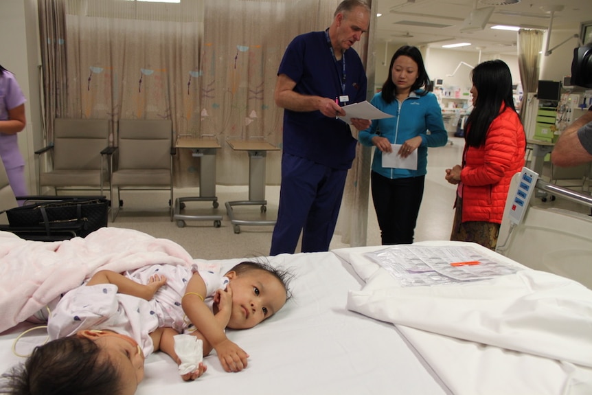 Two twin toddler girls lie on a hospital bed as three adults stand in the background