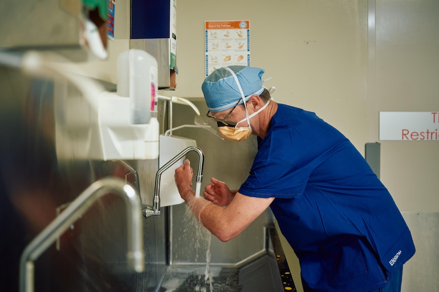 A man in medical scrubs and a cap washing his hands at a sink inside a hospital room.
