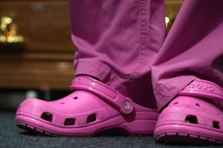 A close up of a pair of legs in pink scrubs and a pair of plastic pink Croc shoes on a dark carpet.
