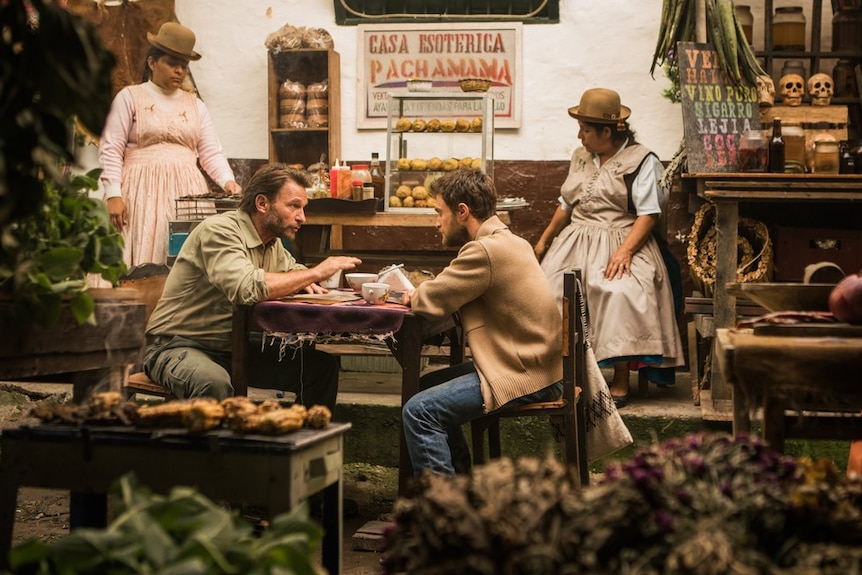 Two characters from the film Jungle sit at an outdoor cafe in South America planning their trip into the Amazon jungle.