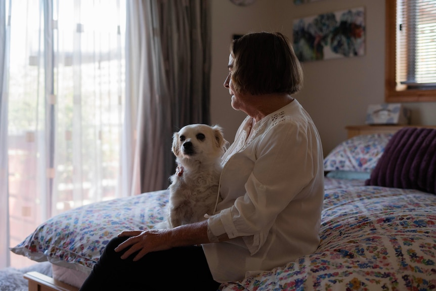 A woman holds a small white dog while sitting on a bed