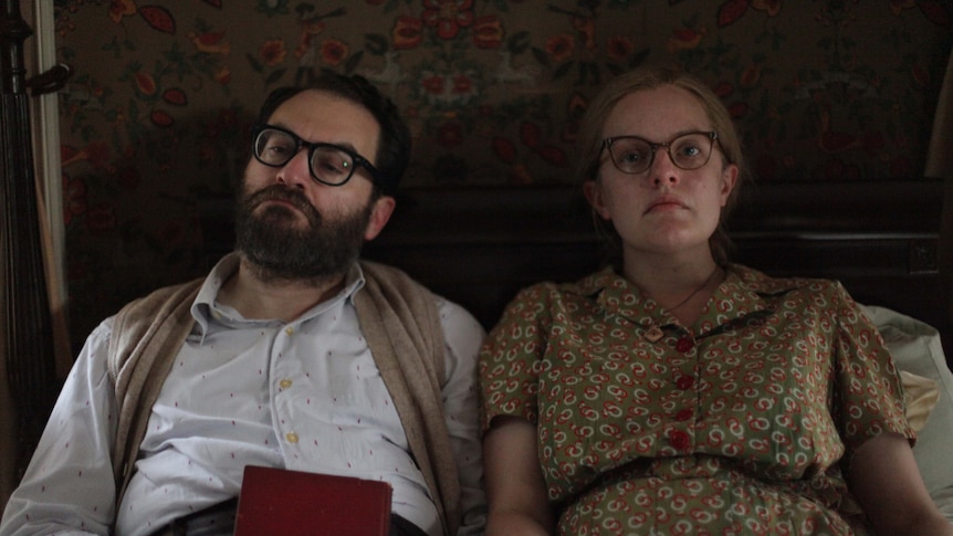 Bespectacled dark hair and bearded man holds red book, sits in bed with bespectacled woman with newspaper in pattern dress.