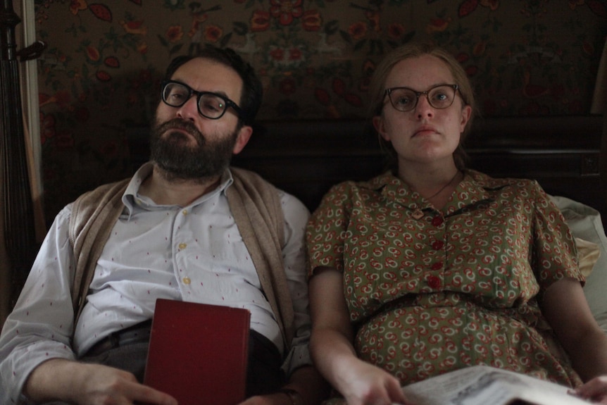 Bespectacled dark hair and bearded man holds red book, sits in bed with bespectacled woman with newspaper in pattern dress.