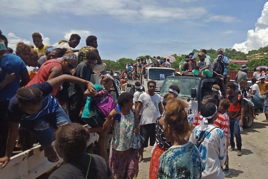 Crowds of people sit on trucks in a rural village.