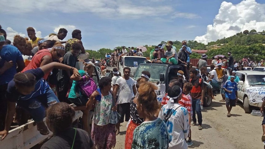 Crowds of people sit on trucks in a rural village.