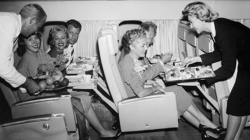 Air cabin crew serve food on trays with passengers seated with their tray tables down.