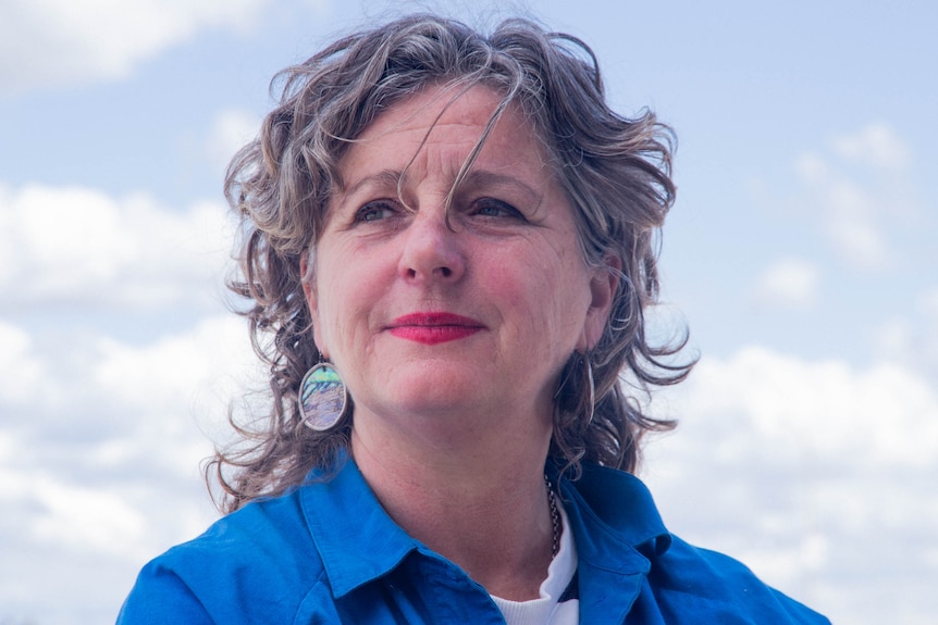 A woman with grey hair and red lipstick looks to left of frame with a slight smile. Clouds in background. 