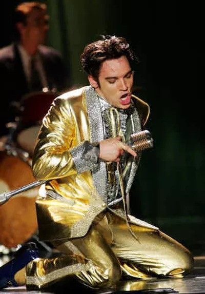 Meyers is on-stage, crouched and singing enthusiastically into a mic, wearing a gold and silver suit.
