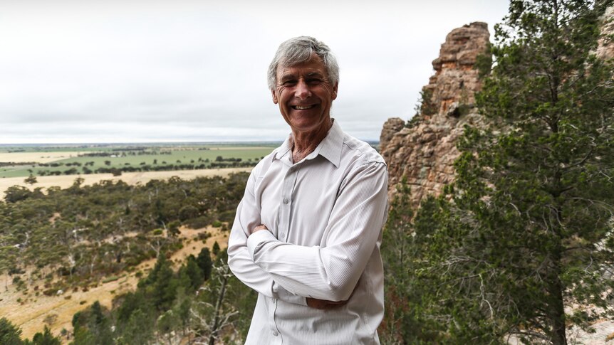 The retired journalist Keith 'Noddy' Lockwood standing on a rock ledge, arms folded,  with the Wimmera plains behind him.