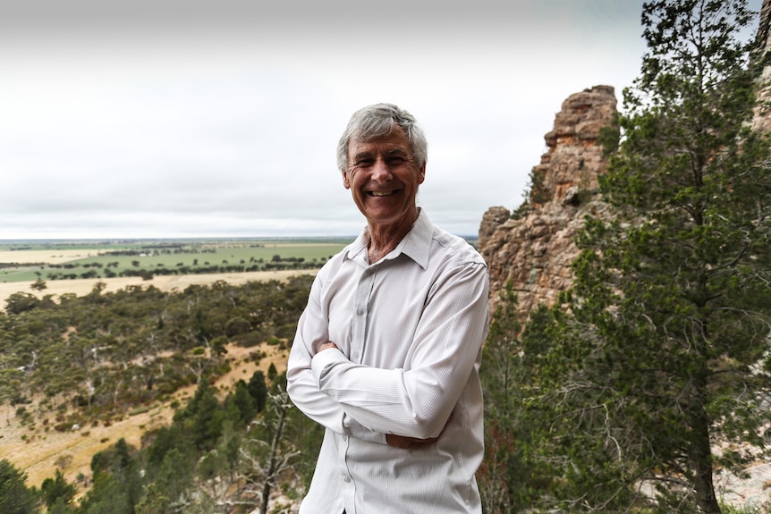 The retired journalist Keith 'Noddy' Lockwood standing on a rock ledge, arms folded,  with the Wimmera plains behind him.