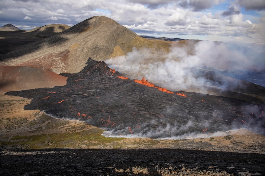 White grey smoke billows from a mass of black rock and lava spreading across a valley. Green hills rise in the background