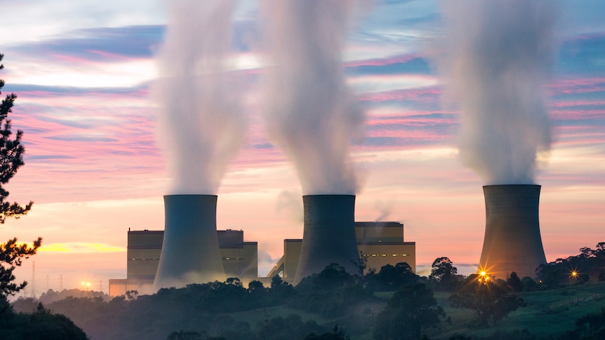 The silhouette of a power station is seen against the sky in the early evening, with thick smoke billowing from three towers.