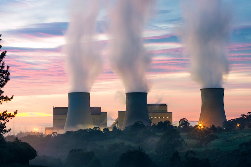 The silhouette of a power station is seen against the sky in the early evening, with thick smoke billowing from three towers.