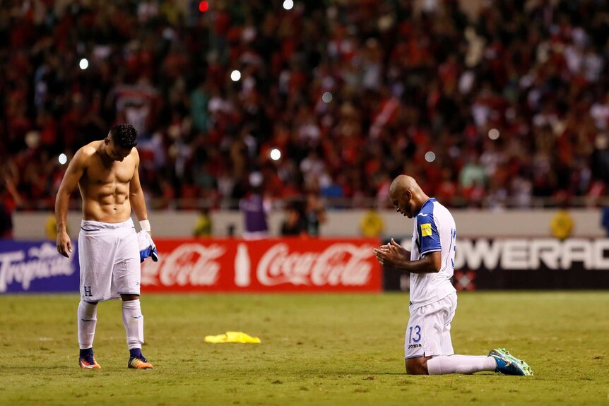 One Honduran player with his shirt of on the pitch and another on his knees praying.