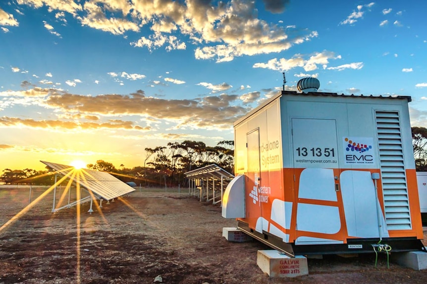 A large shed on raised tinder blocks with Western Power branding, in front of solar panels with the sun setting in background.