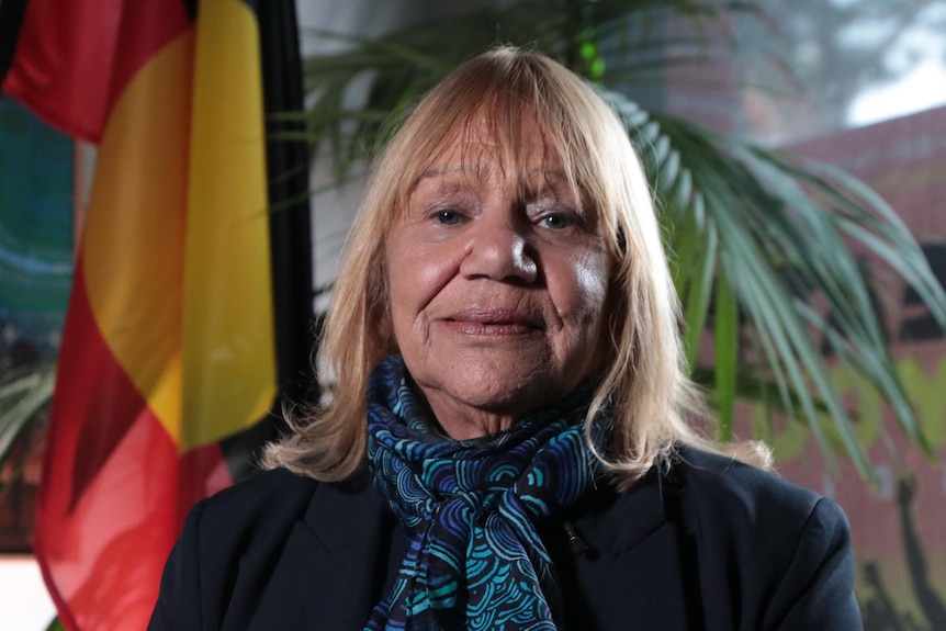 an aboriginal woman wearing a colourful scarf with an aboriginal flag behind her.