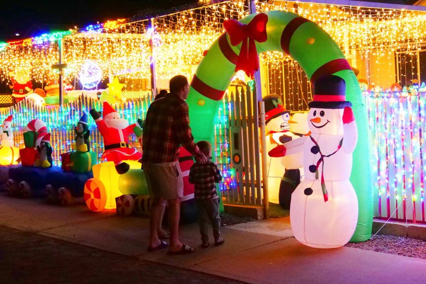 A man, woman and child walk through an inflatable green Christmas wreath into a decorated front yard.