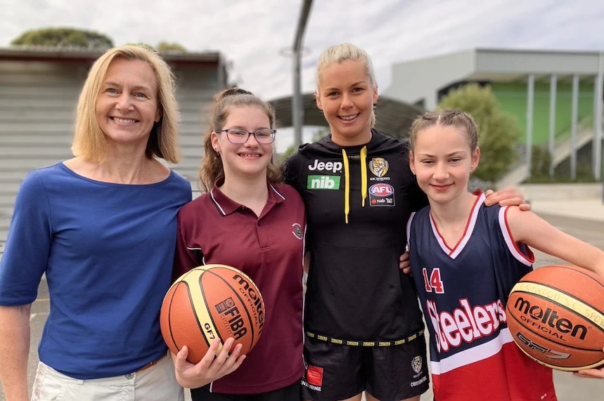 La académica Clare Hanlon y tres mujeres jóvenes que visten diferentes uniformes deportivos en una cancha de baloncesto