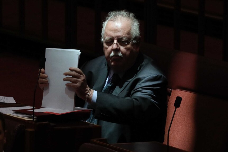 Barry O'Sullivan, bathed in shadow, shuffles papers on his desk in the Senate. He is frowning.