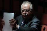 Barry O'Sullivan, bathed in shadow, shuffles papers on his desk in the Senate. He is frowning.