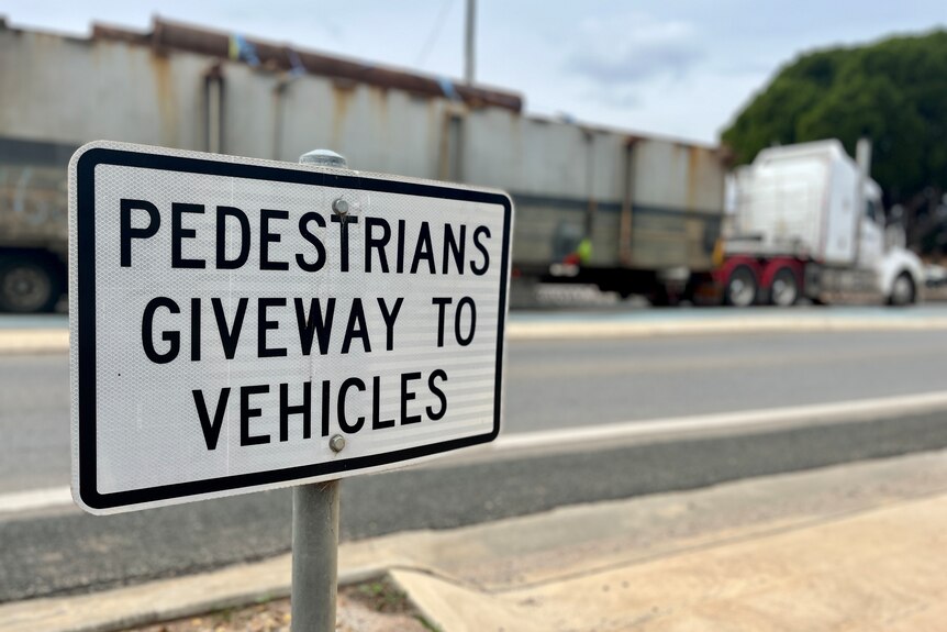 a traffic sign says pedestrians giveway to vehicles at the edge of a crossing. a B double truck in the background.