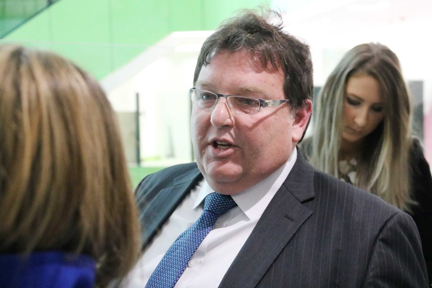 Head and shoulders shot of Lindsay Albonico standing wearing a suit and spectacles talking to reporters.