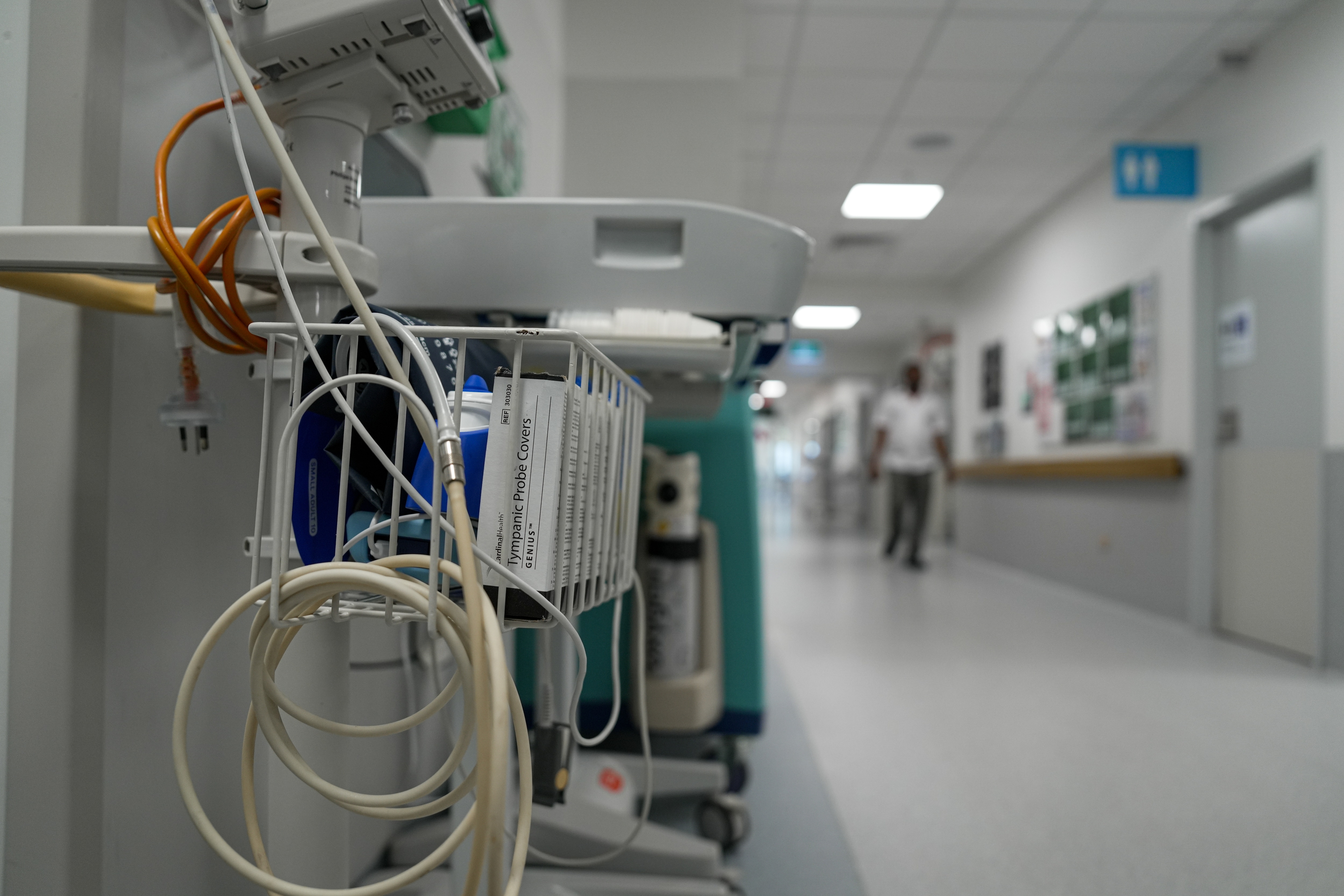 Un carrito lleno de equipo hospitalario se encuentra en un pasillo completamente blanco, con una figura vestida de blanco visible al fondo.
