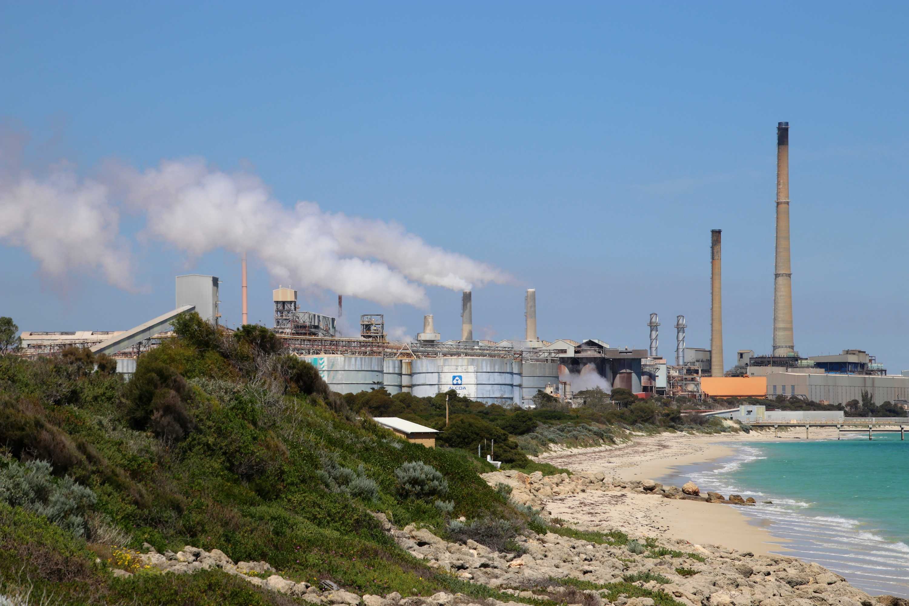 An empty beach leads to a heavy industrial area with large smoke plumes