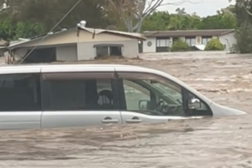 A silver van up to the windows is brown floodwater. A submerged house is half underwater in the background.