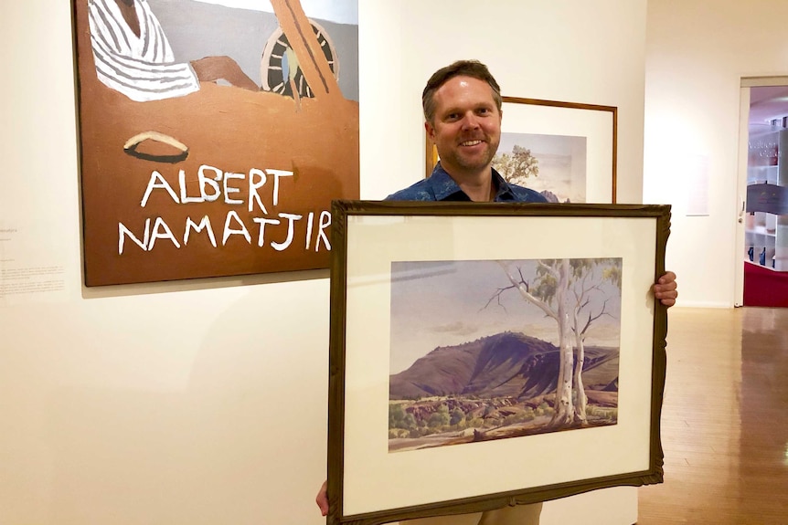 Man standing in gallery holding Mount Hermannsburg painting
