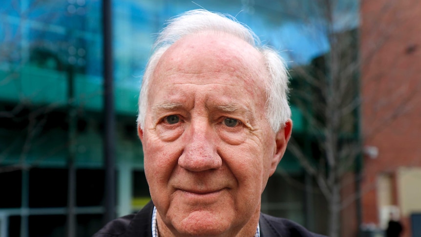 An old white haired man sits at a table outside in a courtyard at the RMIT