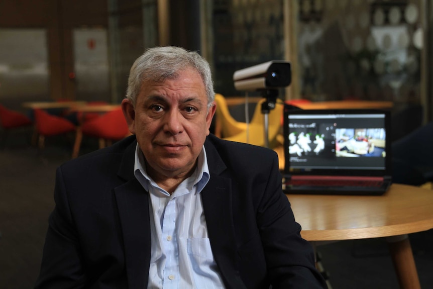 Rustom Kanga in a light blue shirt and black jacket, with a camera and computer on the table behind him