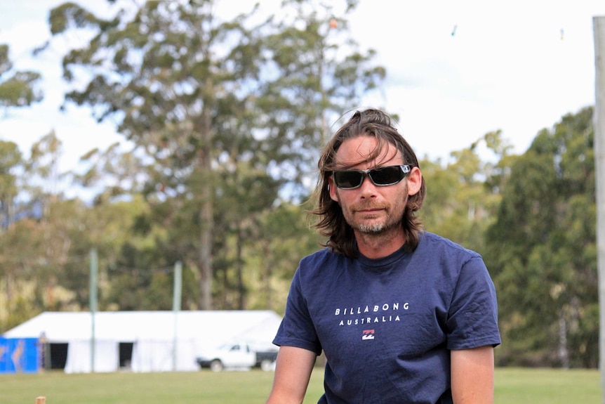 A man wearing a blue tshirt with dark sunglasses on in a bush setting