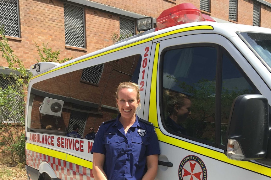 A paramedic smiles as she stands with her hands together leaning against an ambulance.