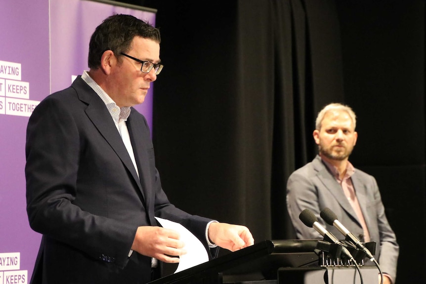 Premier Daniel Andrews stands at a podium addressing the media with Chief Health Officer Brett Sutton to the side.